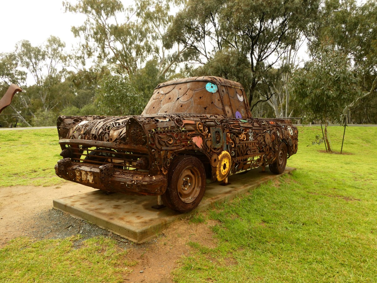 A weathered sculpture of a ute welded from scrap metal, gears and machine parts, standing on a plinth on grass among gum trees.