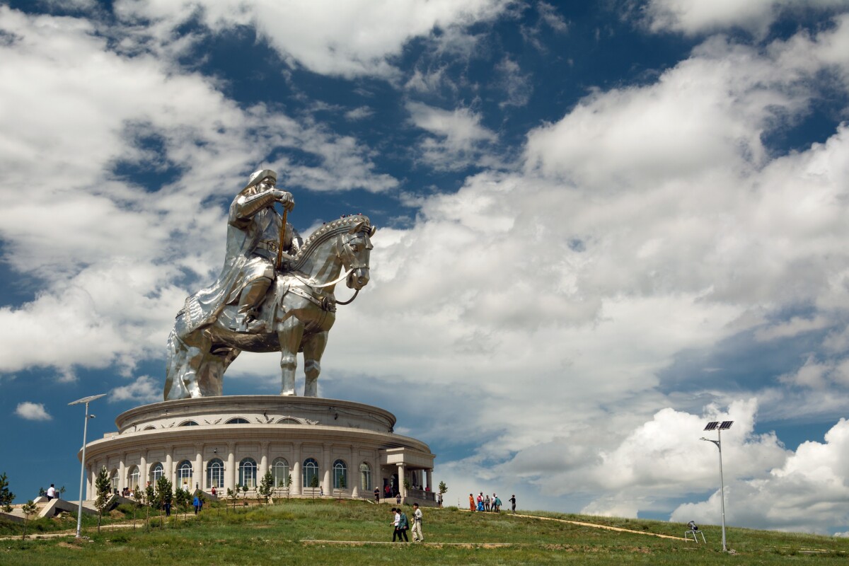 A towering stainless steel statue of Genghis Khan on horseback rising from its round white visitor centre at Tsonjin Boldog, seen under a partly cloudy sky with visitors walking near its base.
