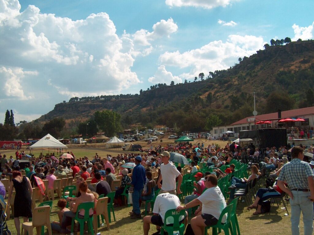 A large crowd seated in rows of plastic chairs on a grass field at a Ficksburg Cherry Festival event, with a rocky hill and event tents behind them.