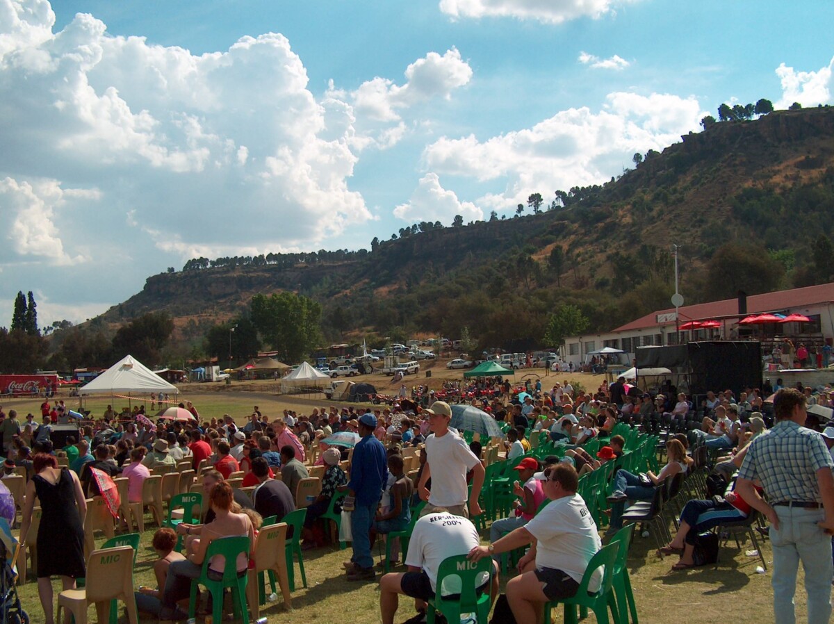 A large crowd seated in rows of plastic chairs on a grass field at a Ficksburg Cherry Festival event, with a rocky hill and event tents behind them.