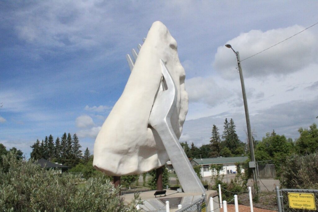 The giant white pyrogy sculpture in Glendon, Alberta, speared by its large fork, on its pedestal under a partly cloudy sky.