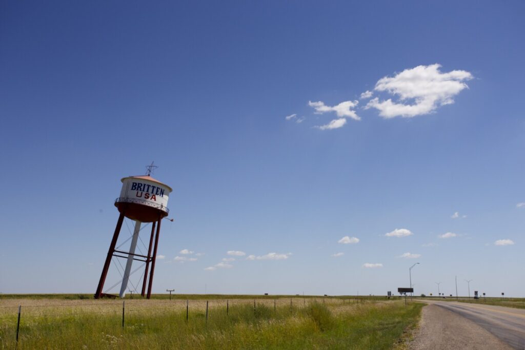 The Britten USA water tower leaning at a sharp angle in a grassy field beside a Texas Panhandle road, under a wide blue sky.