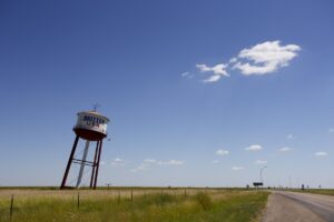 The Britten USA water tower leaning at a sharp angle in a grassy field beside a Texas Panhandle road, under a wide blue sky.