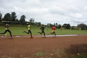 Four runners in training kit stride along a red dirt track in Iten, with sheep grazing in the field behind them.