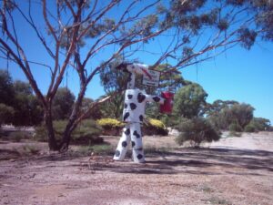 A tin horse painted with black and white cow spots stands beside the Tin Horse Highway road sign under a gum tree.