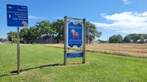 A blue highway sign reading Valkommen, Lindsborg, Little Sweden USA, painted with a red Dala horse, beside a road sign pointing to the pool, museum and downtown.
