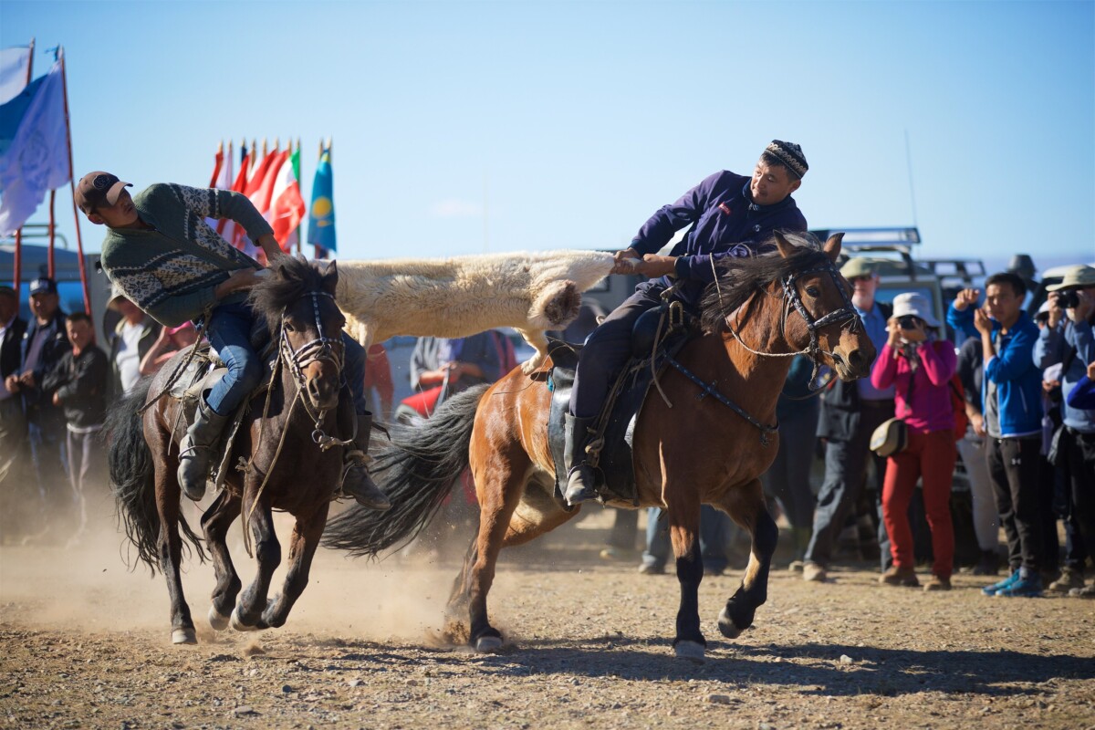 Two horsemen at the Ölgii festival pull a sheepskin between them during a mounted kukbar contest, as a crowd of spectators watches.