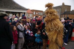A performer in a tall straw costume, the Whittlesey straw bear, led through a Cambridgeshire street by a rope, with musicians following behind.